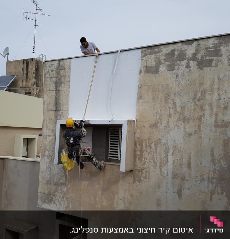 עובד בגובה צובע קיר חיצוני בעזרת חבלים